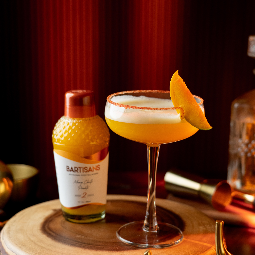  Cocktail with a salt rim and mango wedge next to a bottle of BARTISANS on a wooden surface. 