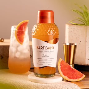 Bartisans Paloma cocktail mixer bottle with a glass of cocktail and grapefruit slices on a table.
