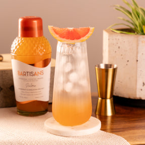 Bartisans Paloma cocktail mixer with a glass of Paloma cocktail on a table with a plant in the background