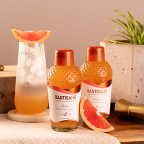 Two bottles of Bartisans cocktail mixers with a cocktail and grapefruit on a wooden surface.