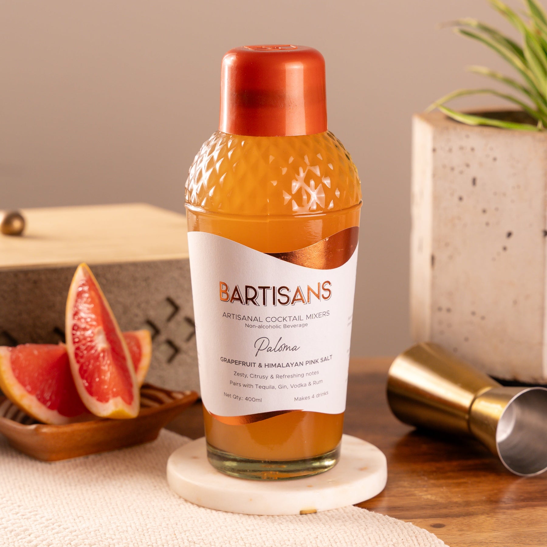 Bottle of BARTISANS Paloma cocktail mixer on a coaster with a background of fruit and decor.