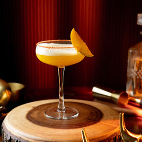 Cocktail with a mango wedge on a wooden coaster with a dark background