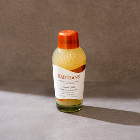Bottle of BARTISANS cocktail mixer on a gray surface with a brown background