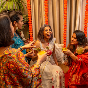 Women celebrating together at an Indian festive gathering, dressed in colorful traditional sarees and holding vibrant cocktails, surrounded by orange marigold garlands and warm festive décor.