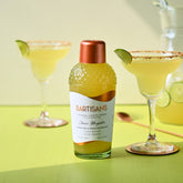 A photograph of a classic margarita made using Bartisans cocktail mixer, displayed on a lime green background. The margarita has a bright yellow color and is garnished with a wedge of lime on the rim of the glass. The cocktail is served in a clear glass with a salt rim, and the label of the Bartisans mixer bottle can be seen in the foreground. The lighting in the photo emphasizes the brightness of the yellow drink and highlights the texture of the salt on the rim.
