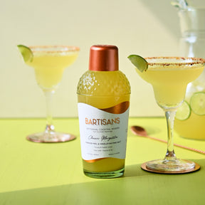 A photograph of a classic margarita made using Bartisans cocktail mixer, displayed on a lime green background. The margarita has a bright yellow color and is garnished with a wedge of lime on the rim of the glass. The cocktail is served in a clear glass with a salt rim, and the label of the Bartisans mixer bottle can be seen in the foreground. The lighting in the photo emphasizes the brightness of the yellow drink and highlights the texture of the salt on the rim.