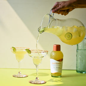 A photograph of a classic margarita being poured into a clear glass from a water jug, with a Bartisans margarita glass bottle visible in the background. The margarita has a pale yellow color and is being poured. The water jug is held by an in-frame hand, and the background features a neutral surface to make the bright colors of the cocktail and glassware stand out. 