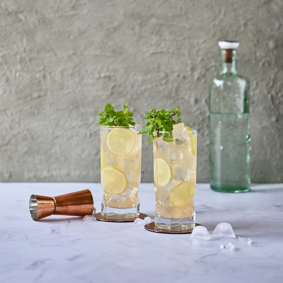 A photograph of two mojitos being prepared and served in clear glasses, with a copper jigger and ice cubes visible below. The mojitos have a pale green color and are garnished with sprigs of fresh mint on top and lime cross sections floating in the drink
