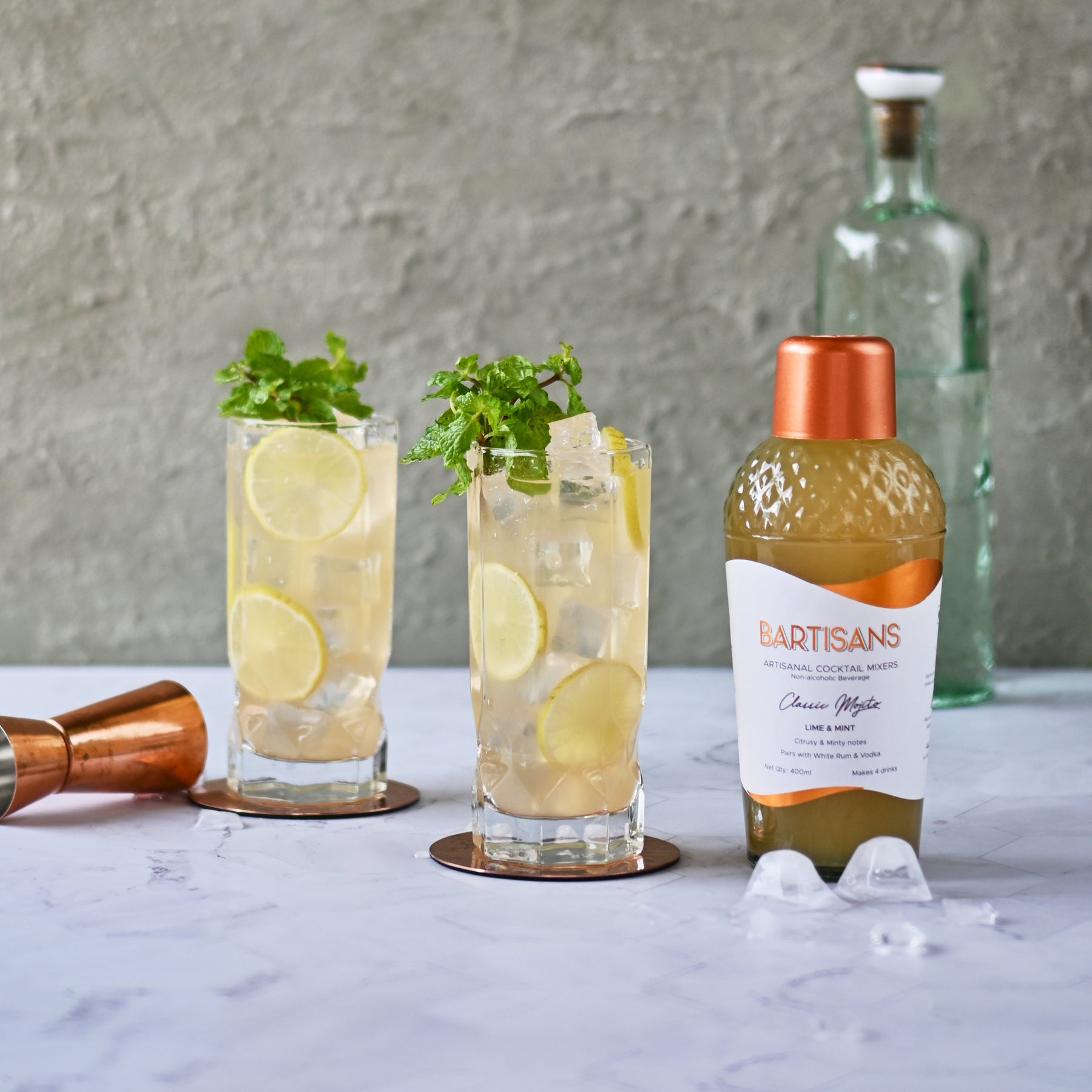 A photograph of two mojitos served in clear glasses, with a bottle of Bartisans mojito cocktail mixer placed beside them. The mojitos have a pale green color and are garnished with sprigs of fresh mint on top. The clear glasses allow the viewer to see the texture and color of the drink, while the Bartisans mojito cocktail mixer bottle provides context for the image and suggests that the cocktails are made using this mixe