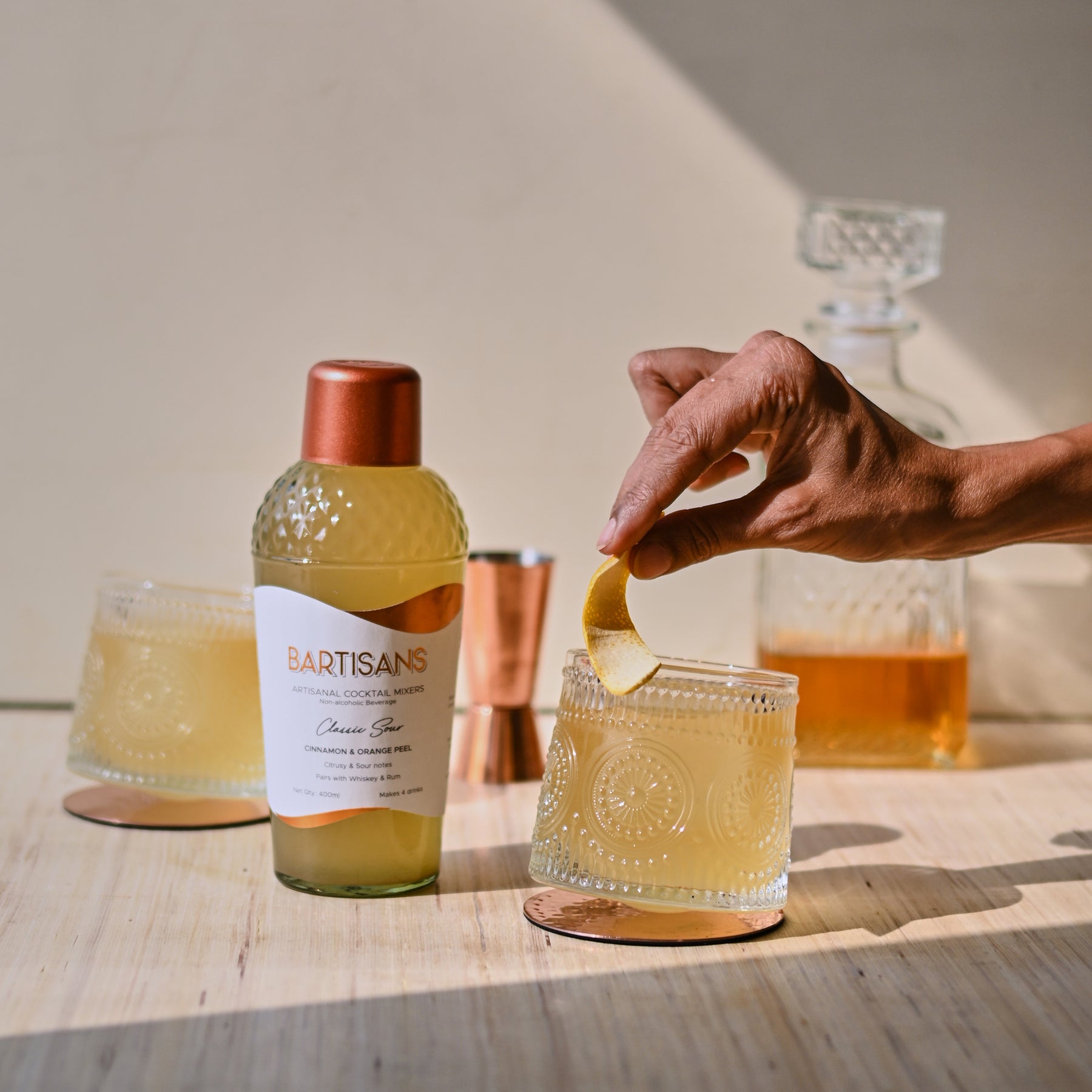 Image of an out of frame hand placing a orange garnish into a glass filled with bartisans classic sour with the mixer bottle kept alongside it