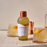 Image of Bartisans classic sour cocktail mixer with two glasses on either side with orange peel floating as garnish and copper jigger behind it